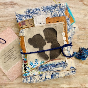Puede incluir: Diario hecho a mano con una cubierta de tela. La cubierta presenta una silueta de una pareja besándose, un patchwork de patrones florales azules y blancos, una borla azul y una pequeña etiqueta rosa con texto.