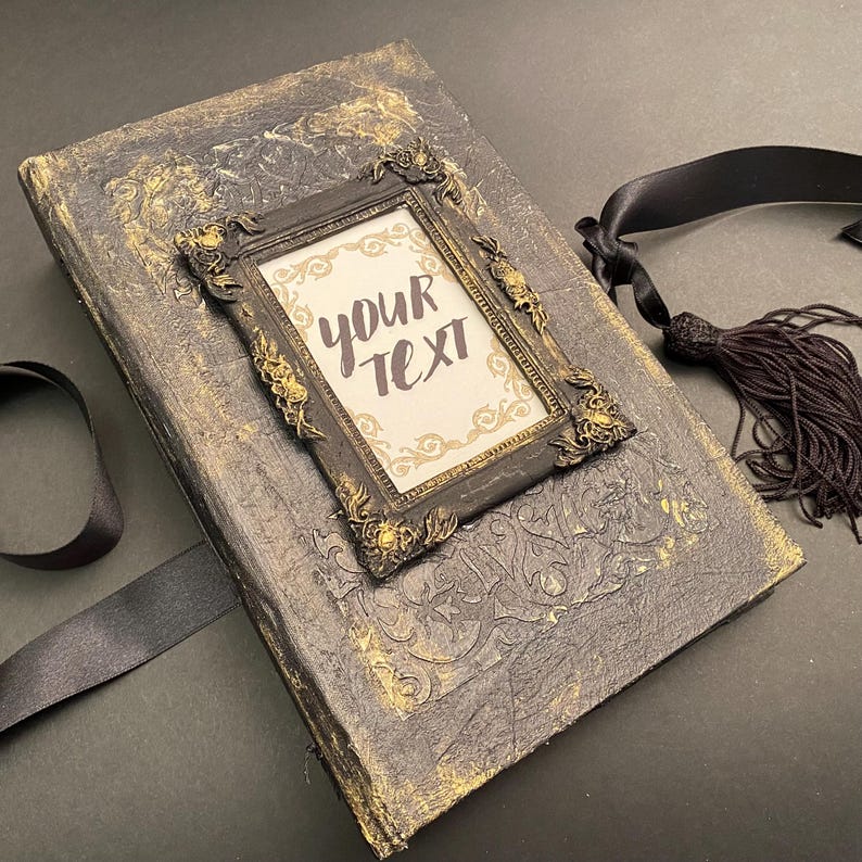 May include: A black and gold journal with an ornate frame on the front. The frame has a white background with the text "your text" written in black. The journal has a black ribbon and tassel.