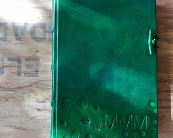 May include: A dark green leather-bound journal with the word "MMMM" embossed on the front cover. The cover is decorated with small flower designs.