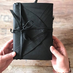 May include: A black leather journal with a unique flap design and tie closure. The journal is held open, showcasing its dark color and textured surface. The cover features a decorative leaf-like pattern.