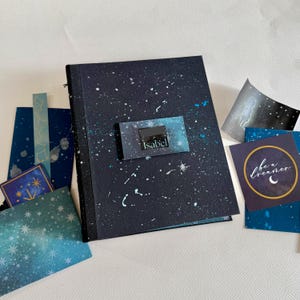 May include: A rectangular notebook with a dark blue cover featuring a white and light blue speckled galaxy pattern. A central rectangular label displays the name Isabel. Several matching stationery cards with celestial designs are arranged around it.