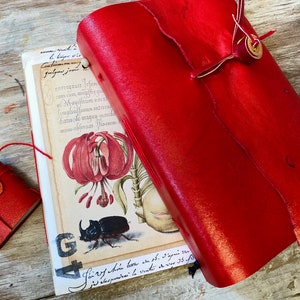 May include: A set of red leather journals and a small matching box. The journals display a botanical illustration of a flower and a beetle. The journals are bound with red thread and have a button closure.