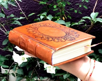 Pode incluir: Um diário de couro castanho com um desenho floral em relevo na capa. O diário tem uma borda decorativa e um marcador. As páginas são brancas e a lombada tem uma textura canelada.