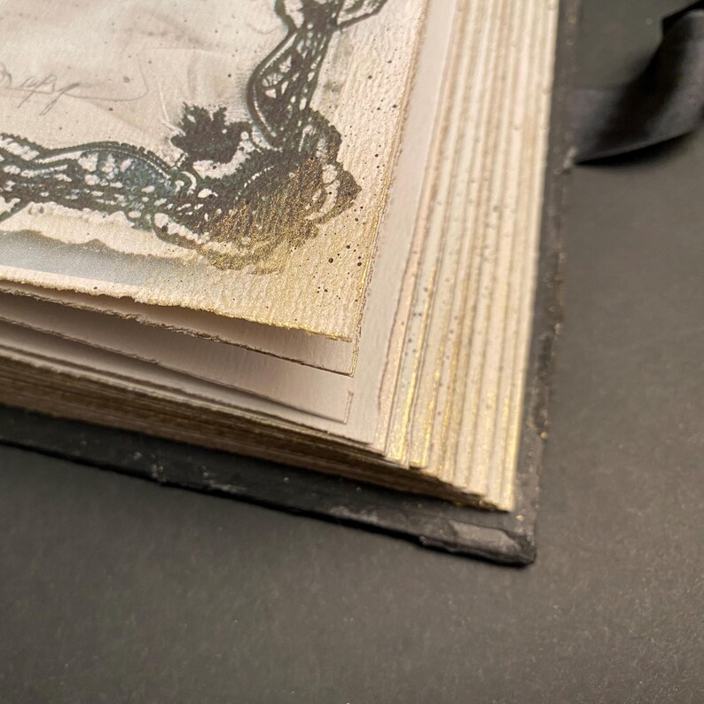 May include: Close-up of the spine of a bound book with gold-gilded edges. The book has a black cover and is made of thick, textured paper.