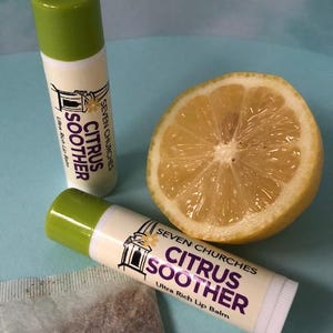 Puede incluir: Dos tubos cilíndricos de bálsamo labial con tapas verdes y etiquetas blancas con el texto Seven Churches Citrus Soother Ultra Rich Lip Balm, colocados junto a un limón amarillo cortado por la mitad y parte de una bolsita de té.
