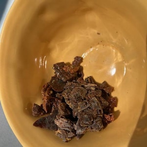 May include: A yellow ceramic bowl filled with irregularly shaped, dark brown and reddish-brown pieces. The bowl has a smooth, glossy finish and is set against a neutral background. The contents appear to be dried or processed food items.