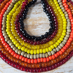 May include: A close-up of a multi-coloured beaded necklace. The necklace is made of small, round beads in shades of red, orange, yellow, and brown. The beads are arranged in a circular pattern, creating a colourful and eye-catching design.