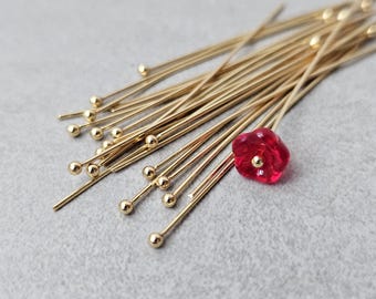 May include: A collection of gold-colored head pins with small ball ends, scattered on a gray surface. One red, flower-shaped bead with a gold center is also present. These pins are likely used for jewelry making or crafting.