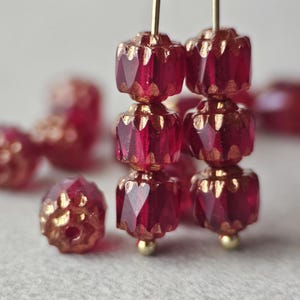May include: A collection of small, faceted glass beads in a deep translucent red color. Each bead features a cylindrical shape with vertical ridges and metallic gold-toned accents along the edges. Two vertical strands of three beads are shown.