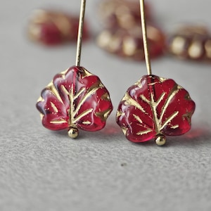 May include: A pair of leaf-shaped earrings. The earrings feature a deep red, translucent glass with gold-colored veins. The leaves are suspended from gold-colored wires and have small gold beads at the base.