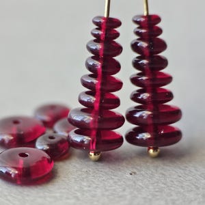 May include: Close-up of two pairs of red glass earrings with gold-colored posts. Each earring is made of stacked, circular beads. Several loose, red glass beads are scattered in the foreground. The earrings and beads are on a light gray surface.