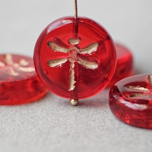 May include: Four red glass beads with a gold dragonfly design. The beads are round and have a smooth, glossy finish.