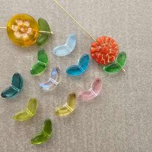 May include: Assortment of colorful glass beads in the shape of leaves. The beads are arranged on a light gray surface. There are two glass flower beads, one yellow and one orange.