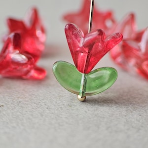 May include: A translucent red glass flower bead with a green glass leaf base, threaded onto a thin metal pin. The bead features a five-petaled blossom shape and a smooth, rounded leaf element at the bottom, with a small metallic stopper at the base.
