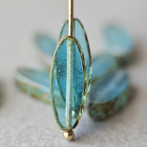 May include: Close-up of oval, light blue glass beads with gold edges and a gold bead at the base. The beads are strung on a gold wire. Other similar beads are blurred in the background. The beads are likely for jewellery making.