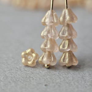 May include: Close-up of delicate, light pink flower-shaped beads with a gold shimmer. Two strands of beads hang vertically, each with four bell-shaped beads. A single flower bead is to the left. The beads are on a gray surface.
