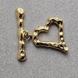 May include: Gold-toned metal heart-shaped toggle clasp with a textured surface. The clasp has a bar that slides through the heart.