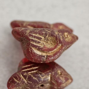 May include: Close-up of several small, red bird-shaped beads. The beads have etched details and a gold-colored finish, with visible wear. The birds are stacked on top of each other, suggesting they are part of a larger piece of jewelry or craft.