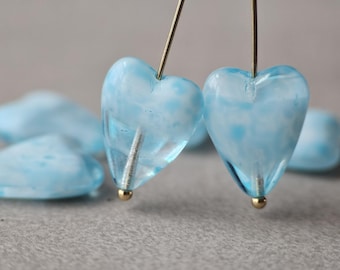 May include: Two light blue heart-shaped glass beads with white marbling are suspended on gold-coloured pins. The beads have a translucent quality, and a few more heart-shaped beads are visible in the background. The beads are likely for jewellery making.