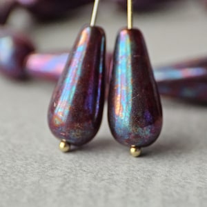May include: Close-up of a pair of iridescent teardrop-shaped earrings. The earrings are a deep burgundy color with hints of blue and green, and have small gold-colored beads at the bottom. The earrings are on gold-colored wires.