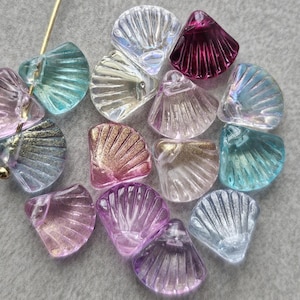 May include: A collection of iridescent seashell-shaped beads in various colors, including pink, purple, teal, and clear. The beads have a textured, fan-like design and are arranged on a gray surface. A gold-colored pin is visible.