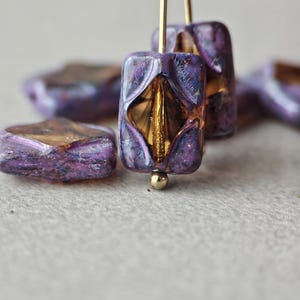 May include: Close-up of several purple and gold beads. The beads have a rectangular shape with a gold interior and a shimmering purple exterior. Some beads are in focus, while others are blurred in the background. The beads are on a light grey surface.