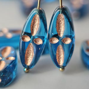May include: Translucent oval-shaped blue glass beads featuring a metallic copper-colored cross pattern on the surface. The beads are arranged in a group, with two beads standing upright on thin metal pins, showcasing the reflective glass texture.