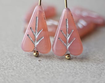 May include: A pair of pink, triangular earrings with a silver leaf design. The earrings have gold-coloured accents at the bottom and are suspended from gold-coloured wires. The background is blurred, focusing on the jewellery.
