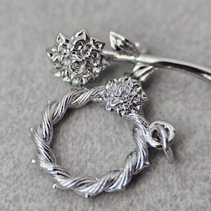 May include: Silver charm featuring a twisted rope design with two flower accents. The flowers are detailed with a raised center and petals.