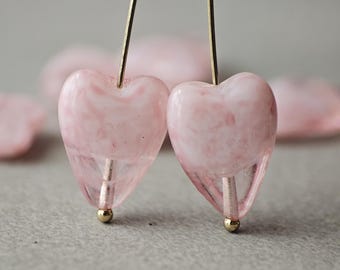 May include: Close-up of two pink heart-shaped beads with gold-coloured accents. The beads are translucent with a marbled effect, suspended on gold-coloured wires. The background is blurred, with additional pink heart shapes.