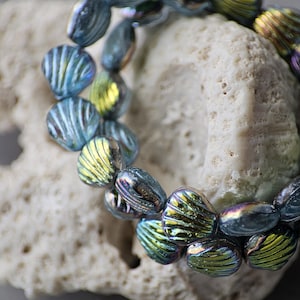 May include: A strand of iridescent blue and green glass beads with a ribbed texture. The beads are teardrop-shaped and are resting on a piece of white coral. The beads have a metallic sheen.