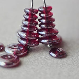 May include: Close-up of handmade earrings featuring stacks of red-toned, disc-shaped beads. The beads have a reflective, metallic sheen. Several loose beads are scattered nearby. The earrings are on a light-colored surface.