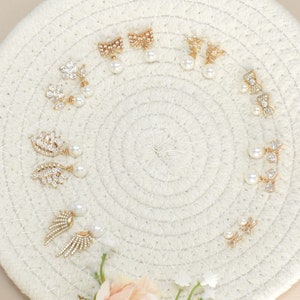 May include: A collection of gold-toned earrings with various designs, including bows, flowers, and leaf shapes, each featuring a dangling faux pearl. The earrings are arranged on a white, woven mat, showcasing their intricate details and elegant style.