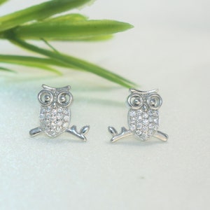 May include: A pair of silver owl stud earrings with clear crystal accents. The owls are perched on a branch.