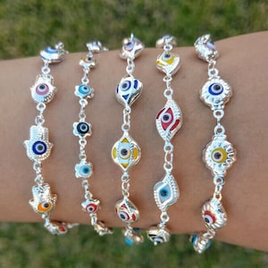 May include: Five silver bracelets with different colored evil eye charms. The charms are in the shape of a hand, a flower, and a teardrop. The bracelets are on a person's wrist.