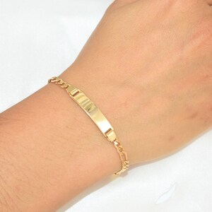 Könnte beinhalten: Ein goldfarbenes ID-Armband mit rechteckiger Platte und Panzerketten-Design. Das Armband wird an einem Handgelenk gezeigt, wobei die Platte zentriert ist und die Kette um das Handgelenk verläuft. Das Armband besteht aus glänzendem, reflektierendem Metall.