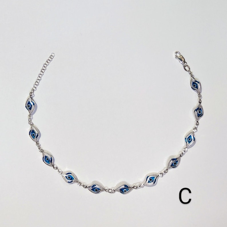 K&ouml;nnte beinhalten: Silberfarbenes Fu&szlig;kettchen mit einer Reihe von blau-wei&szlig;en Evil-Eye-Anh&auml;ngern. Die Anh&auml;nger sind oval und mit einem detaillierten Augendesign versehen. Das Fu&szlig;kettchen hat eine filigrane Kette und einen Verschluss. Der Buchstabe "C" ist unten rechts zu sehen.