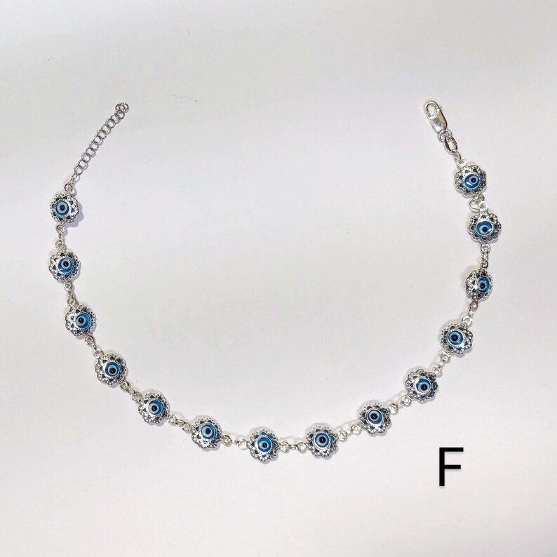 K&ouml;nnte beinhalten: Silberfarbenes Fu&szlig;kettchen mit einem sich wiederholenden Muster aus blau-wei&szlig;en Evil-Eye-Anh&auml;ngern. Die Anh&auml;nger sind in einem dekorativen floralen Design angeordnet. Das Fu&szlig;kettchen hat eine Kettenverl&auml;ngerung f&uuml;r eine verstellbare Gr&ouml;&szlig;e und einen Karabinerverschluss.
