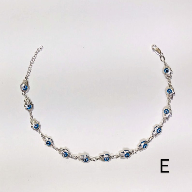 K&ouml;nnte beinhalten: Silberfarbene Halskette mit einem sich wiederholenden Muster aus Hamsa-Hand-Anh&auml;ngern, jeweils mit einem blauen Auge in der Mitte. Die Halskette hat eine zarte Kette und einen Karabinerverschluss. Der Buchstabe "E" befindet sich unten rechts.