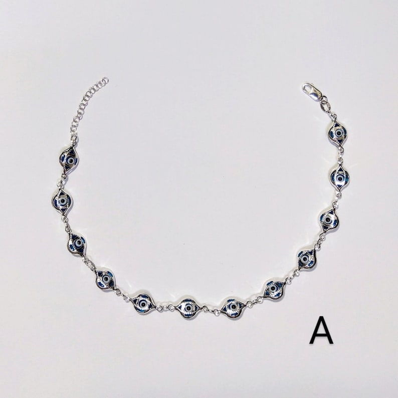 K&ouml;nnte beinhalten: Silberfarbene Halskette mit einer Reihe von blau-wei&szlig;en Evil-Eye-Anh&auml;ngern. Die Anh&auml;nger haben ein detailliertes Augendesign. Die Halskette wird vor wei&szlig;em Hintergrund mit dem Buchstaben "A" gezeigt.