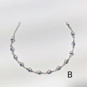 K&ouml;nnte beinhalten: Silberfarbenes Kettenarmband mit einer Reihe von blauen und wei&szlig;en Evil-Eye-Anh&auml;ngern. Das Armband hat ein filigranes Design mit einem Karabinerverschluss und einer verstellbaren Kette. Die Anh&auml;nger sind mit einer blauen Iris und einem wei&szlig;en Hintergrund versehen.