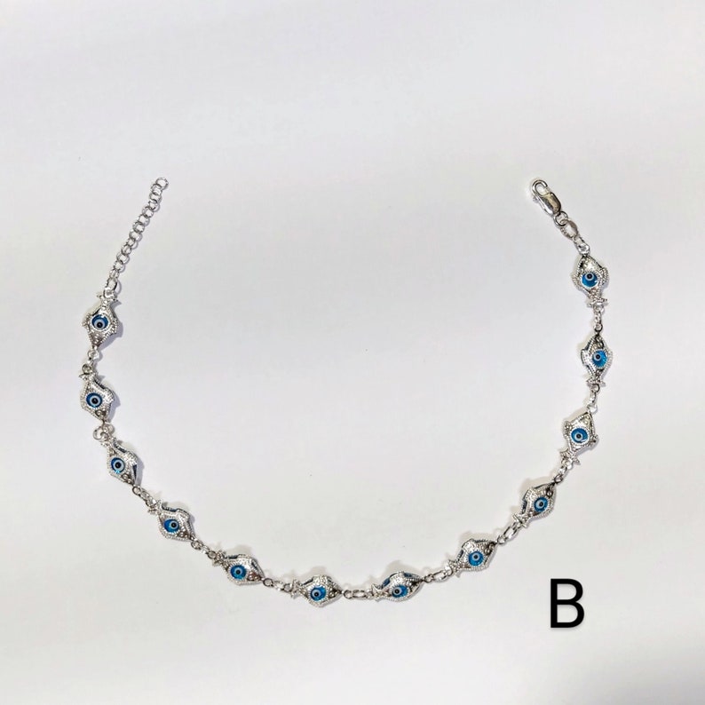 K&ouml;nnte beinhalten: Silberfarbenes Kettenarmband mit einer Reihe von blauen und wei&szlig;en Evil-Eye-Anh&auml;ngern. Das Armband hat ein filigranes Design mit einem Karabinerverschluss und einer verstellbaren Kette. Die Anh&auml;nger sind mit einer blauen Iris und einem wei&szlig;en Hintergrund versehen.