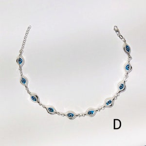K&ouml;nnte beinhalten: Ein silberfarbenes Armband mit einer Reihe von blauen und wei&szlig;en Evil-Eye-Anh&auml;ngern. Die Anh&auml;nger sind oval und haben eine blaue Iris und eine wei&szlig;e Pupille. Das Armband liegt auf wei&szlig;em Hintergrund. Der Buchstabe "D" ist zu sehen.