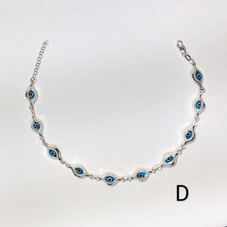 K&ouml;nnte beinhalten: Ein silberfarbenes Armband mit einer Reihe von blauen und wei&szlig;en Evil-Eye-Anh&auml;ngern. Die Anh&auml;nger sind oval und haben eine blaue Iris und eine wei&szlig;e Pupille. Das Armband liegt auf wei&szlig;em Hintergrund. Der Buchstabe "D" ist zu sehen.