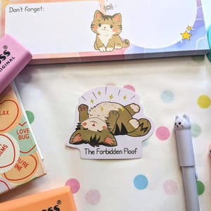 May include: A die-cut sticker featuring a brown tabby cat lying on its back with its paws up, surrounded by yellow sparkles. The text below the cat reads The Forbidden Floof in black font. The sticker is surrounded by stationery items.