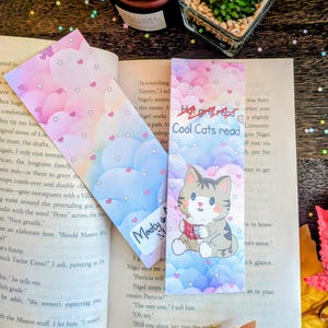 May include: Two rectangular paper bookmarks featuring a pastel gradient background of clouds, small pink hearts, and white stars. One bookmark displays a cartoon tabby cat reading a book with the text Cool Cats read and Hot girls read.