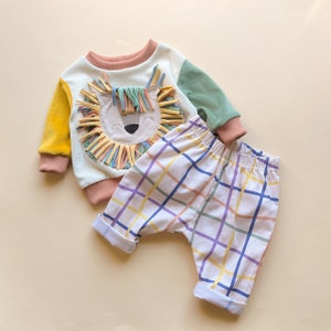 May include: A white baby sweater with a colorful lion applique and a pair of white and multi-colored plaid pants.