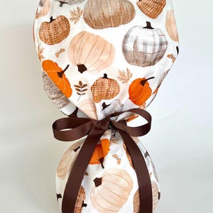 May include: A scrub cap featuring a white background with a repeating pattern of pumpkins in shades of orange, beige, and grey, accented with small brown leaves and acorns. The cap is gathered at the base and tied with a dark brown grosgrain ribbon.