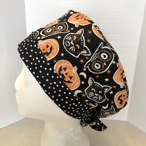 May include: A scrub cap featuring a black background with a repeating pattern of orange jack-o'-lanterns, black cats, and small stars. The cap includes a wide, folded brim band in black fabric with a white polka dot pattern and tie-back ends.