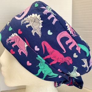 May include: A blue surgical scrub hat with a pattern of pink, green, and white dinosaurs and pink hearts.